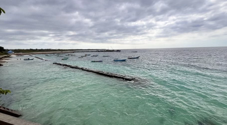 Waikelo Beach features clear blue-green water with numerous small boats floating near a long wooden pier under a cloudy sky.