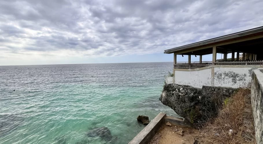 A white building with an open-air terrace sits on a rocky cliff overlooking turquoise and dark blue ocean water under a cloudy sky.