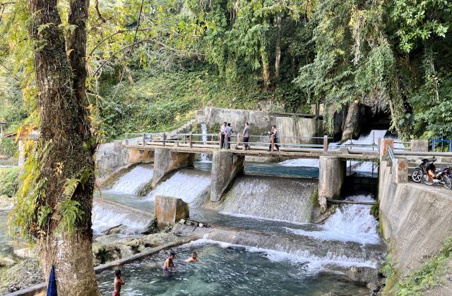The Waikelo Sawah Dam features concrete spillways with cascading water, surrounded by dense green jungle, as people walk on a bridge and boys swim below.