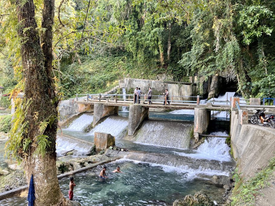 The Waikelo Sawah Dam features concrete spillways with cascading water, surrounded by dense green jungle, as people walk on a bridge and boys swim below.