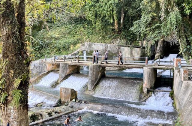 The Waikelo Sawah Dam features concrete spillways with cascading water, surrounded by dense green jungle, as people walk on a bridge and boys swim below.