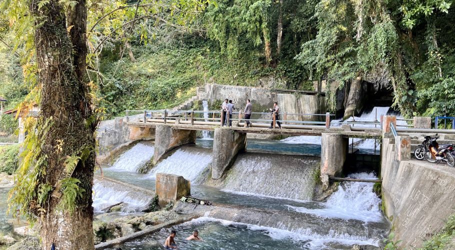 The Waikelo Sawah Dam features concrete spillways with cascading water, surrounded by dense green jungle, as people walk on a bridge and boys swim below.
