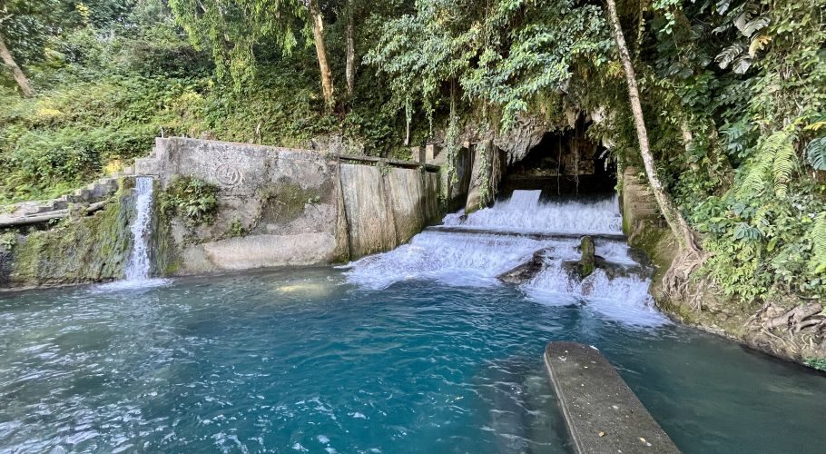 A concrete dam structure with several waterfalls cascades into a bright blue-green pool, surrounded by dense green tropical vegetation.