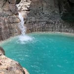 A powerful white waterfall plunges into a vibrant turquoise pool, surrounded by dark, layered rock cliffs in a natural grotto.