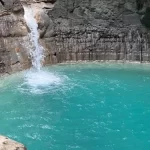 A powerful white waterfall plunges into a vibrant turquoise pool, surrounded by dark, layered rock cliffs in a natural grotto.