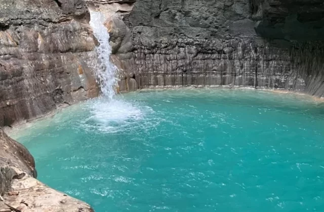 A powerful white waterfall plunges into a vibrant turquoise pool, surrounded by dark, layered rock cliffs in a natural grotto.