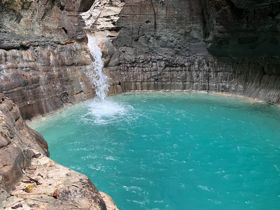 A powerful white waterfall plunges into a vibrant turquoise pool, surrounded by dark, layered rock cliffs in a natural grotto.