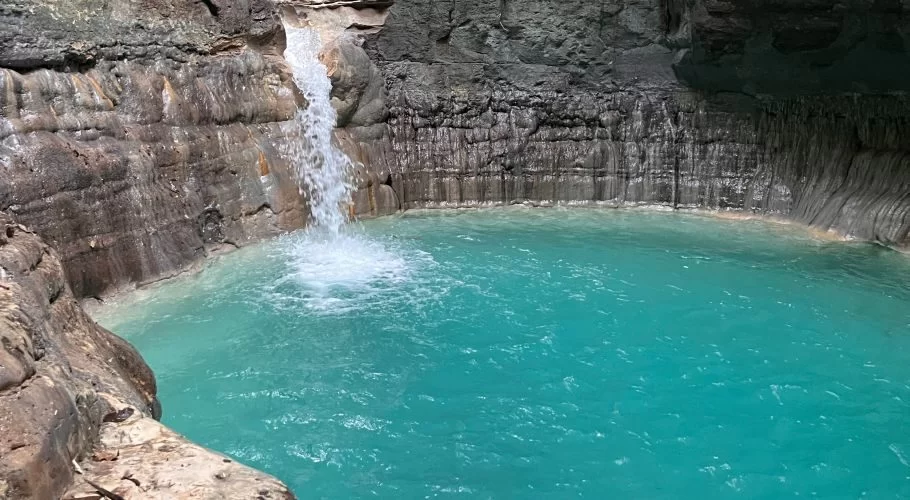 A powerful white waterfall plunges into a vibrant turquoise pool, surrounded by dark, layered rock cliffs in a natural grotto.