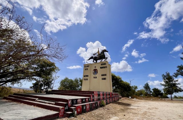 A bronze statue of a spear-wielding rider on a rearing horse stands atop a beige monument under a bright blue sky with white clouds, flanked by dry trees.