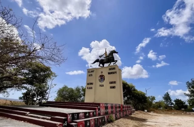 A bronze statue of a spear-wielding rider on a rearing horse stands atop a beige monument under a bright blue sky with white clouds, flanked by dry trees.