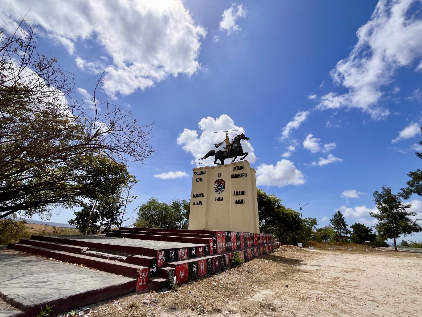 A bronze statue of a spear-wielding rider on a rearing horse stands atop a beige monument under a bright blue sky with white clouds, flanked by dry trees.