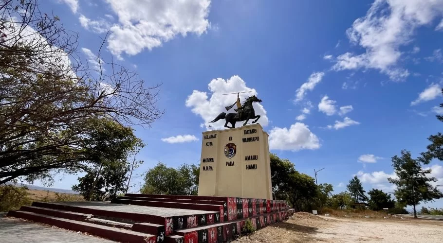 A bronze statue of a spear-wielding rider on a rearing horse stands atop a beige monument under a bright blue sky with white clouds, flanked by dry trees.