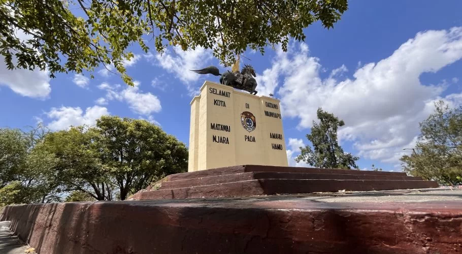 A light yellow monument topped with a dark statue of a horseback rider holding a spear stands under a bright blue sky with white clouds, framed by green trees.