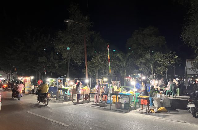 Brightly lit food stalls, including grilled corn vendors, line a dark street at Waingapu City Park at night, with people and motorbikes.