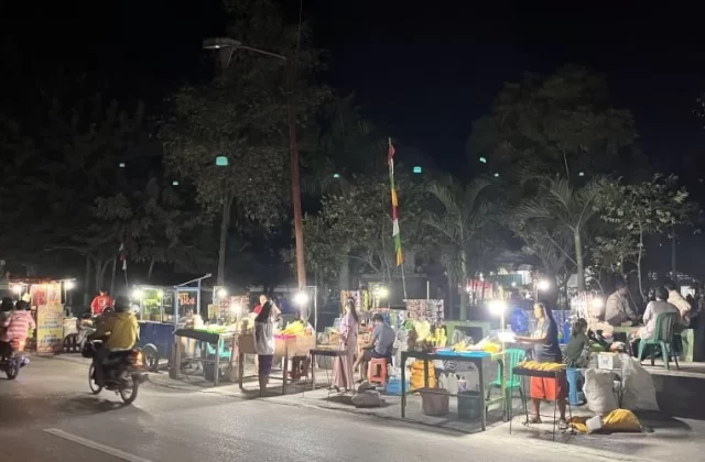 Brightly lit food stalls, including grilled corn vendors, line a dark street at Waingapu City Park at night, with people and motorbikes.