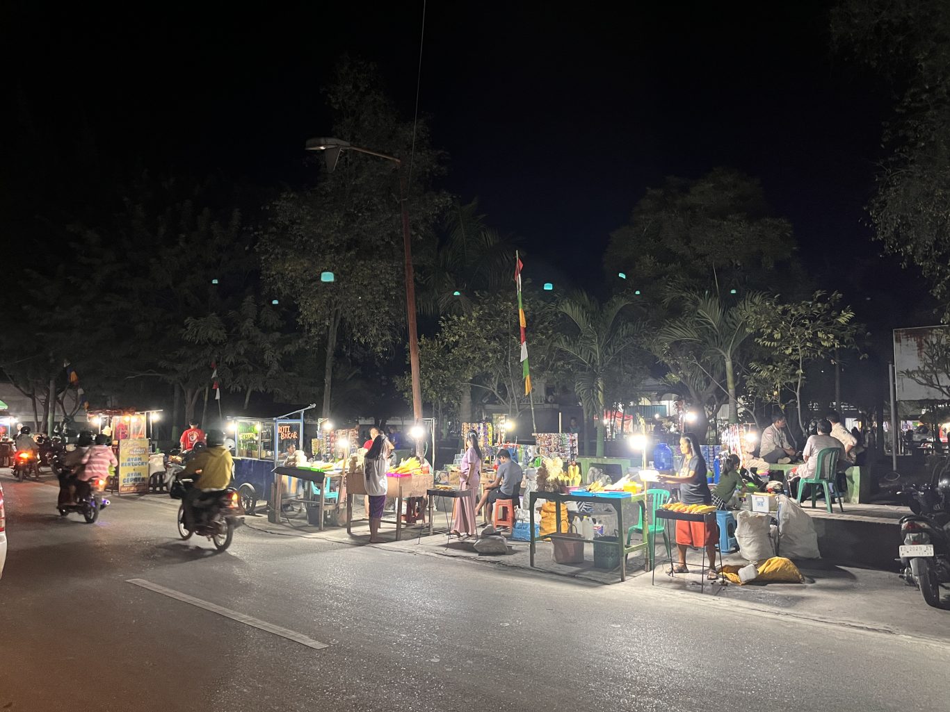 Brightly lit food stalls, including grilled corn vendors, line a dark street at Waingapu City Park at night, with people and motorbikes.