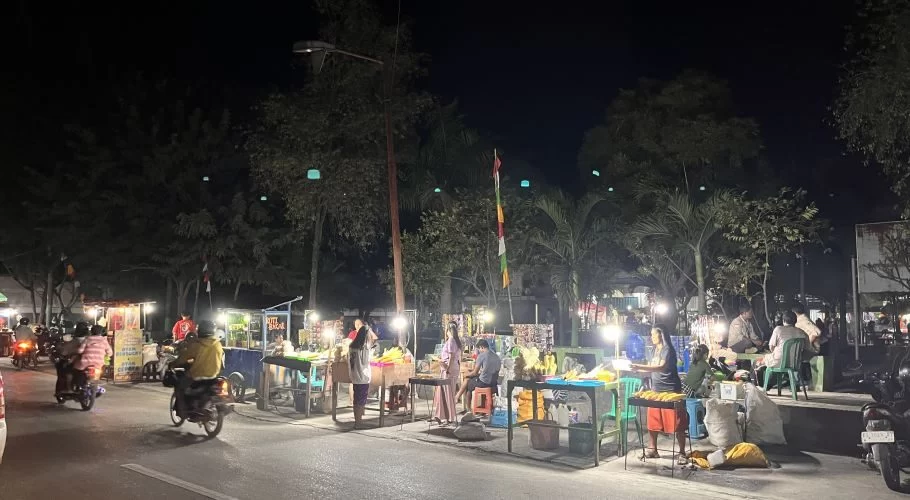 Brightly lit food stalls, including grilled corn vendors, line a dark street at Waingapu City Park at night, with people and motorbikes.