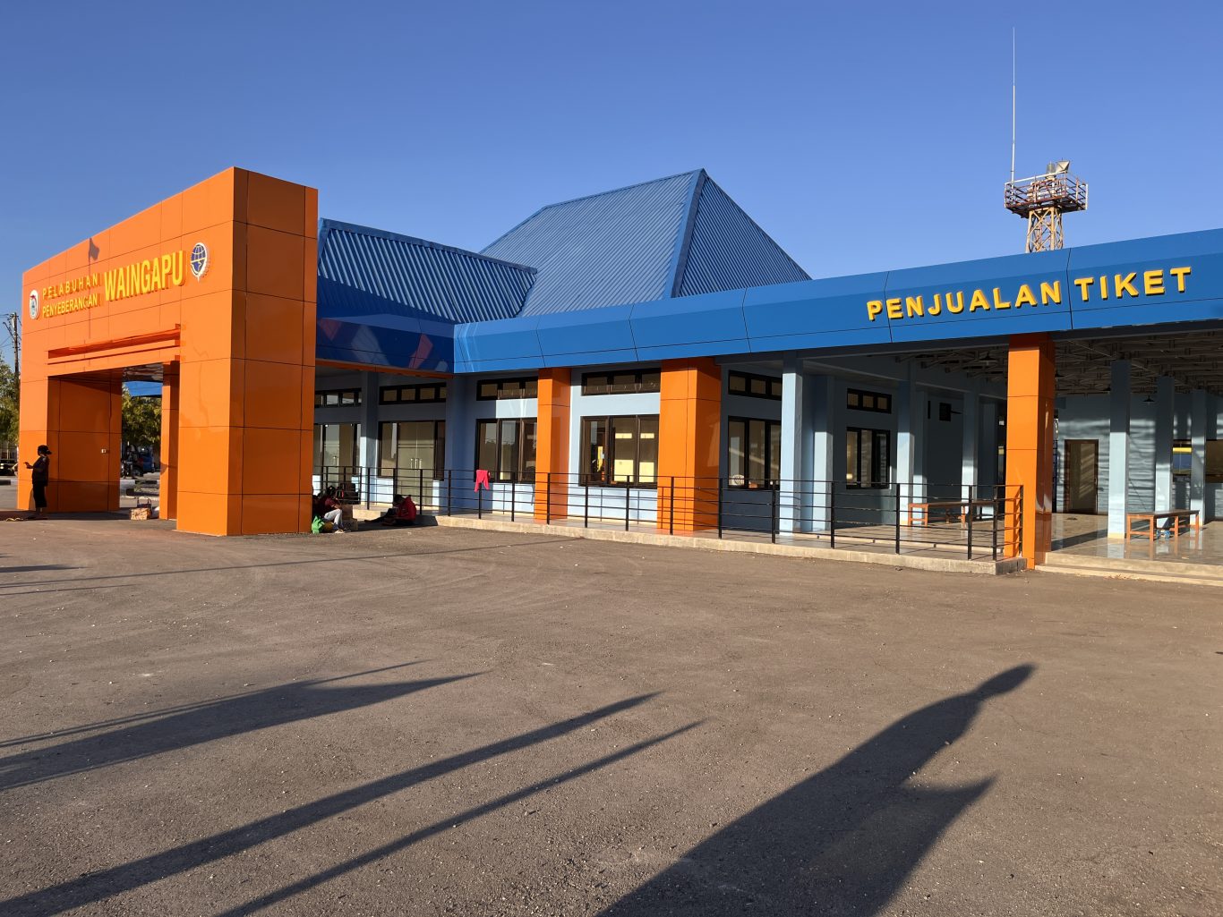 The modern Waingapu Ferry Port terminal building features a bright orange entrance arch and a blue-roofed ticket sales area under a clear sky.