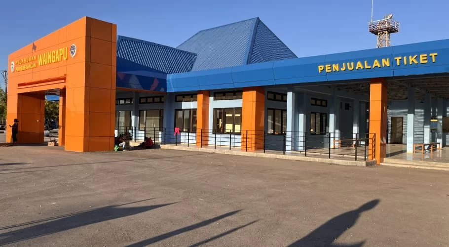 The modern Waingapu Ferry Port terminal building features a bright orange entrance arch and a blue-roofed ticket sales area under a clear sky.