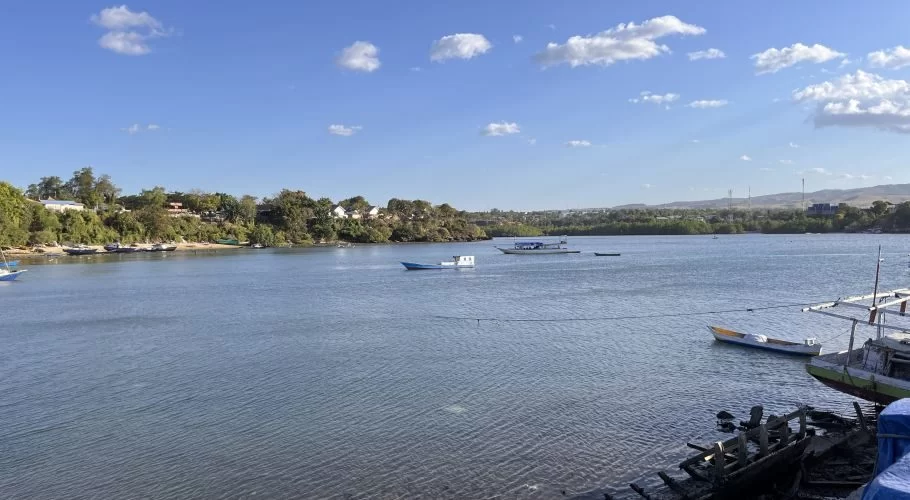 Small fishing boats dot the calm blue waters of Waingapu Old Port, with a lush green shoreline and distant hills under a bright blue sky.