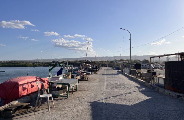 The concrete pier of Waingapu Old Port is lined with small fishing boats and market stalls under a bright blue sky, with distant green hills.