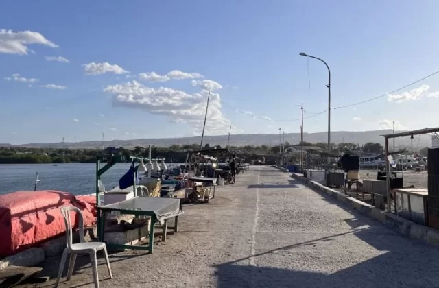 The concrete pier of Waingapu Old Port is lined with small fishing boats and market stalls under a bright blue sky, with distant green hills.