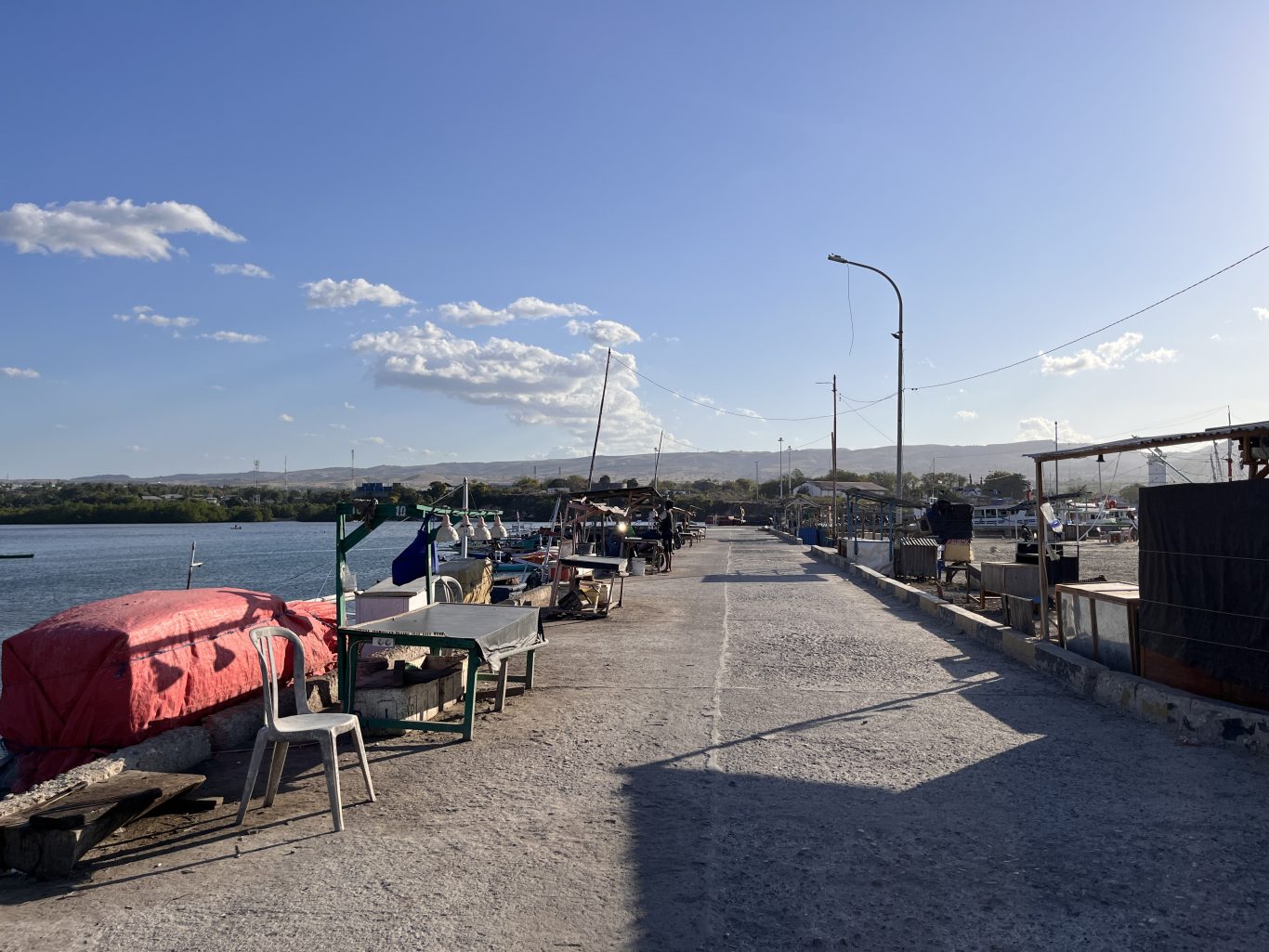 The concrete pier of Waingapu Old Port is lined with small fishing boats and market stalls under a bright blue sky, with distant green hills.