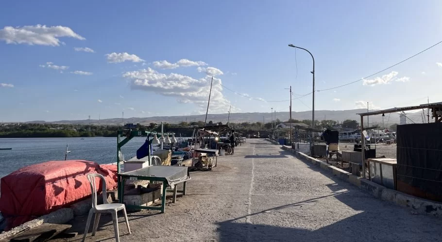 The concrete pier of Waingapu Old Port is lined with small fishing boats and market stalls under a bright blue sky, with distant green hills.