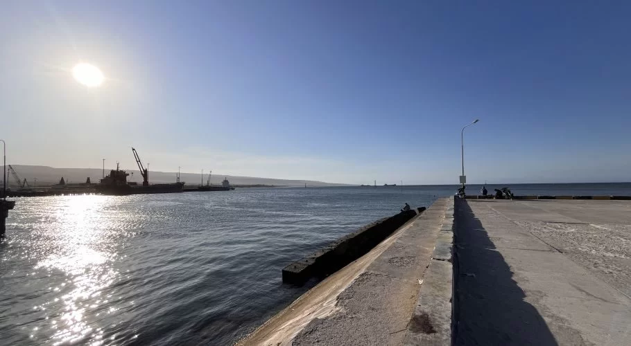 Waingapu Old Port under a bright blue sky, showing a concrete pier, sun-glared water, and silhouetted cargo ships against hills.