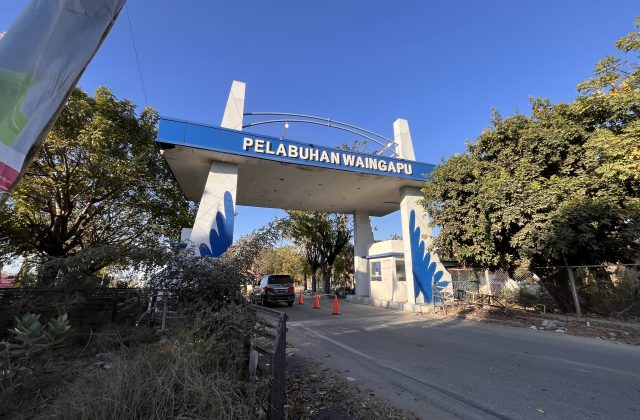 The blue and white entrance gate of Pelabuhan Waingapu stands under a clear blue sky, flanked by lush green trees, with a car entering the port.