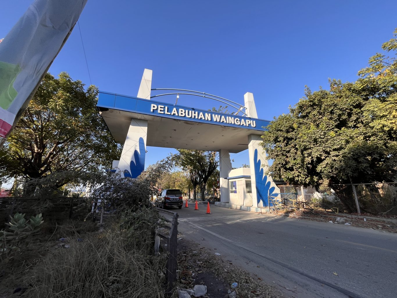 The blue and white entrance gate of Pelabuhan Waingapu stands under a clear blue sky, flanked by lush green trees, with a car entering the port.