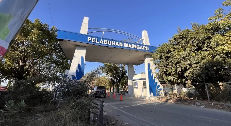 The blue and white entrance gate of Pelabuhan Waingapu stands under a clear blue sky, flanked by lush green trees, with a car entering the port.