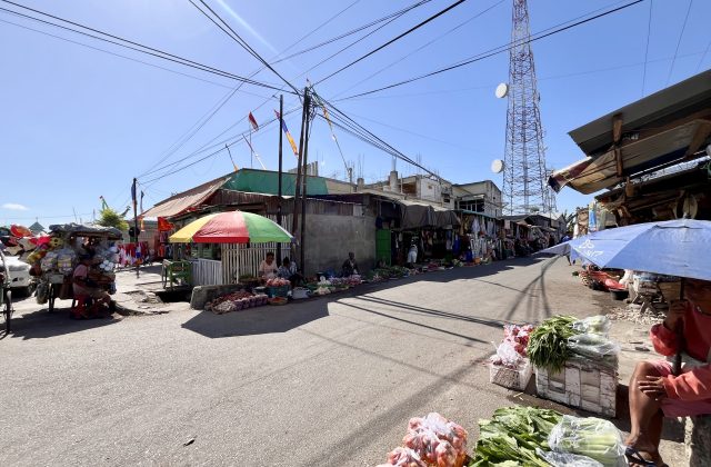 A sunny street at Waingapu Traditional Market shows vendors selling fresh produce under colorful umbrellas, with buildings and a cell tower.