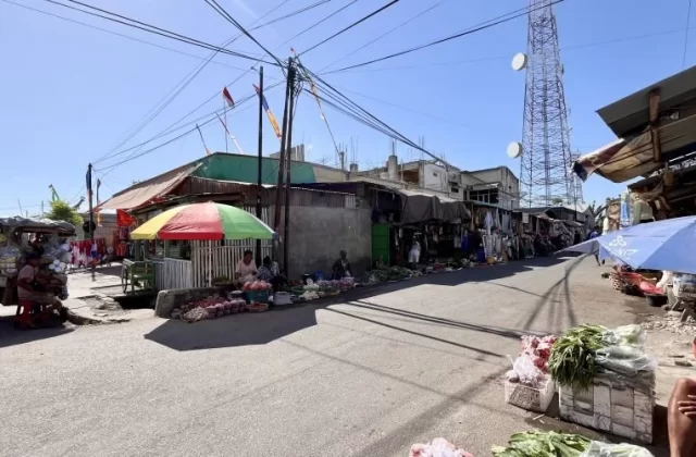A sunny street at Waingapu Traditional Market shows vendors selling fresh produce under colorful umbrellas, with buildings and a cell tower.