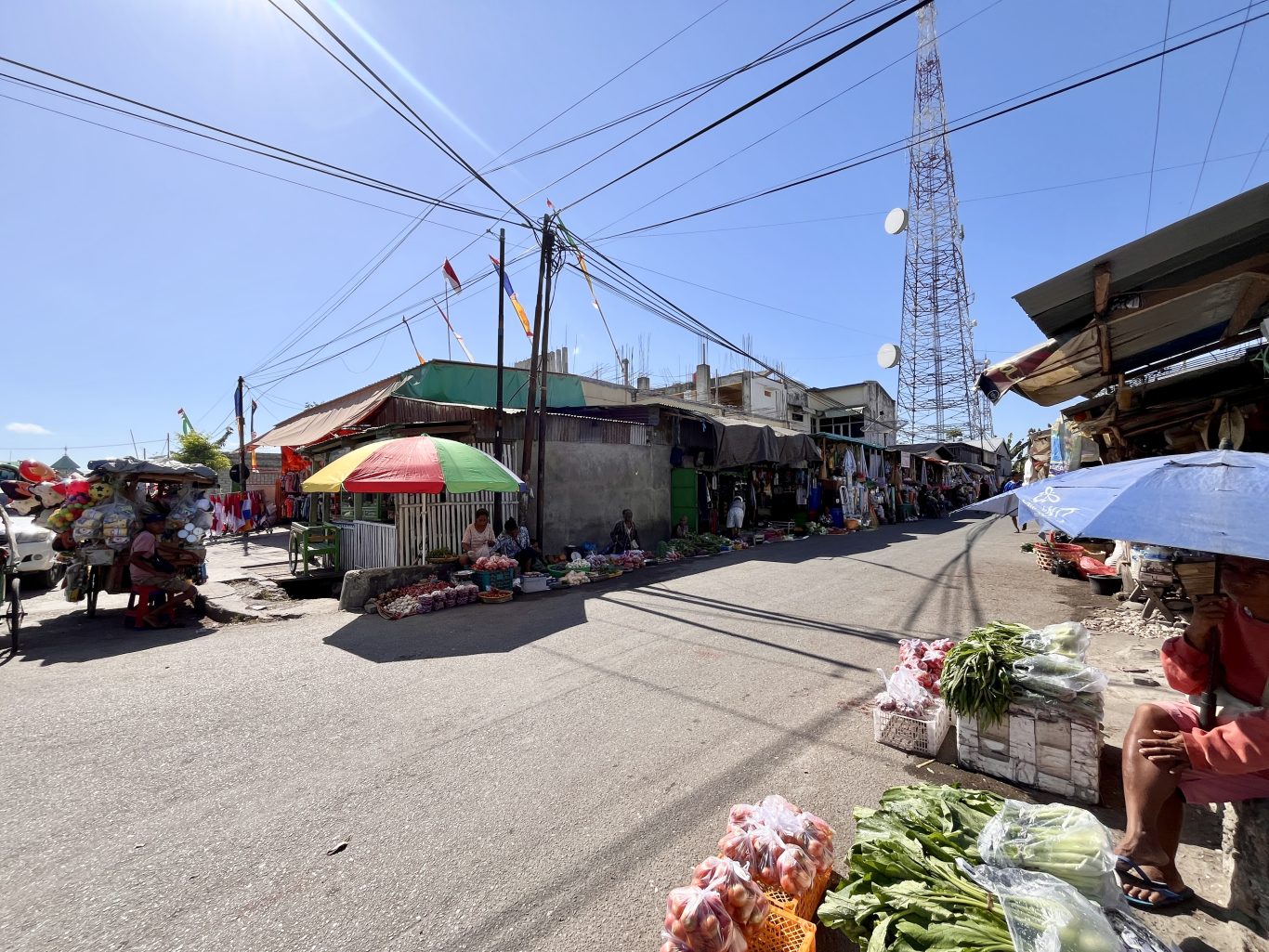 A sunny street at Waingapu Traditional Market shows vendors selling fresh produce under colorful umbrellas, with buildings and a cell tower.