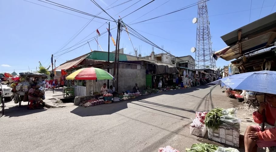 A sunny street at Waingapu Traditional Market shows vendors selling fresh produce under colorful umbrellas, with buildings and a cell tower.