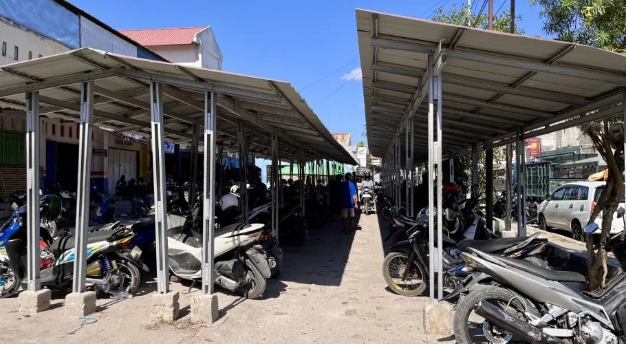 A sunny outdoor scene shows two long, grey metal covered parking structures filled with many motorbikes, with a few people walking between them.