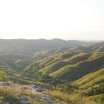 Undulating golden-brown savanna hills with scattered green trees stretch under a clear blue sky, illuminated by soft, warm sunlight.