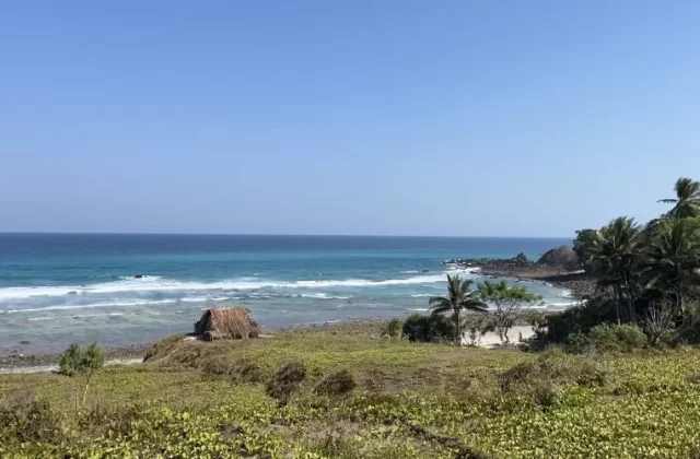 A clear blue sky overlooks a tranquil turquoise ocean with white waves breaking on Waiwuang Beach, featuring a small thatched hut, green vegetation, and palm trees.