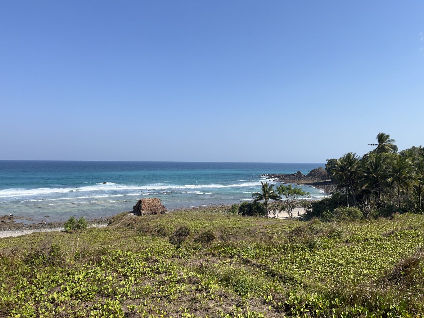 A clear blue sky overlooks a tranquil turquoise ocean with white waves breaking on Waiwuang Beach, featuring a small thatched hut, green vegetation, and palm trees.