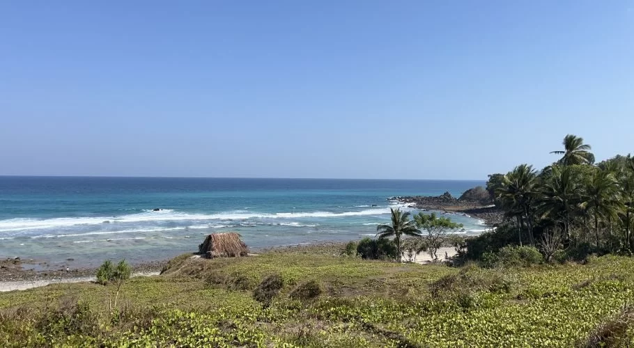 A clear blue sky overlooks a tranquil turquoise ocean with white waves breaking on Waiwuang Beach, featuring a small thatched hut, green vegetation, and palm trees.