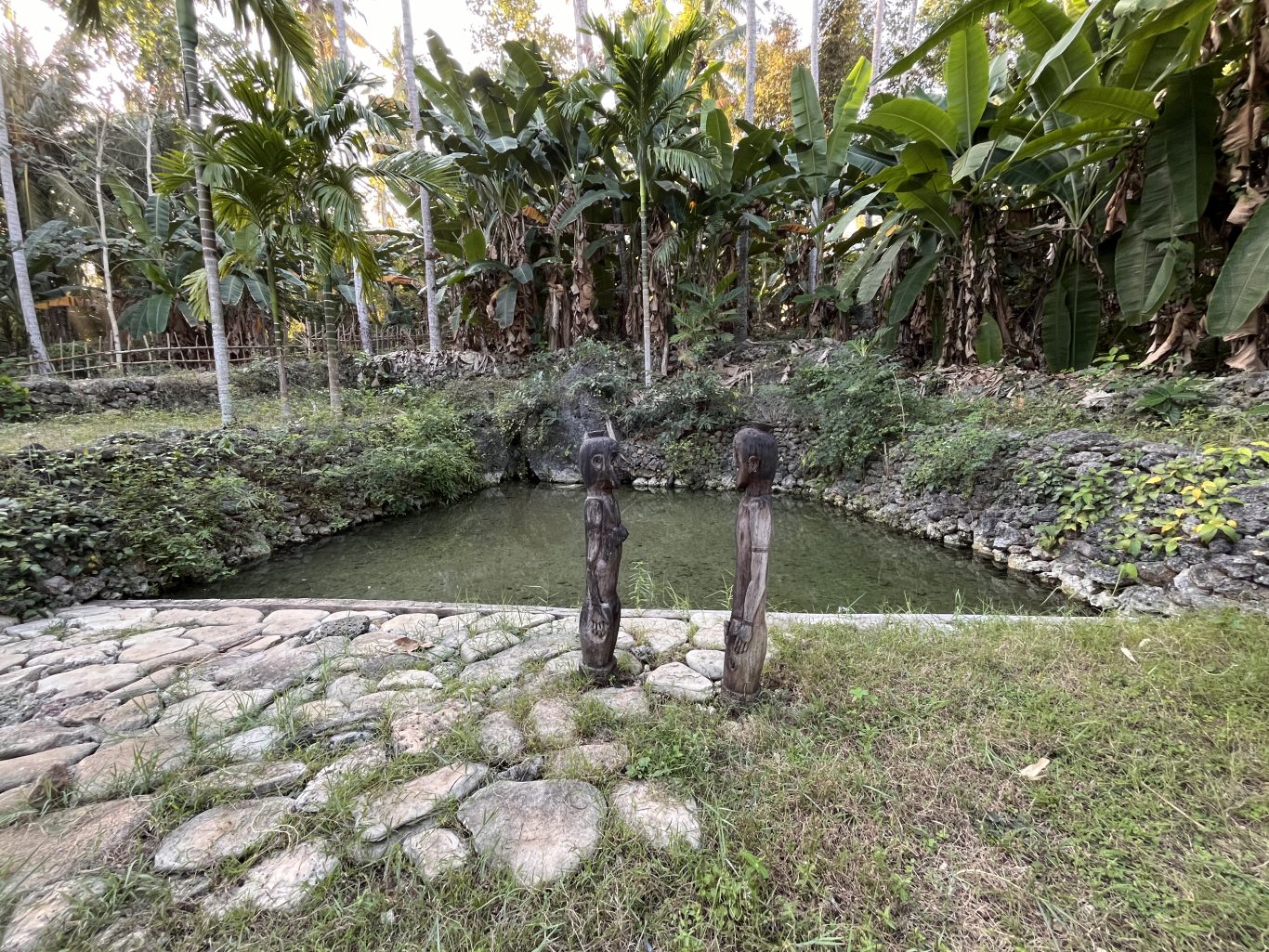 Two dark wooden statues of a male and female stand on a stone path beside a clear spring pool, surrounded by lush green tropical foliage.