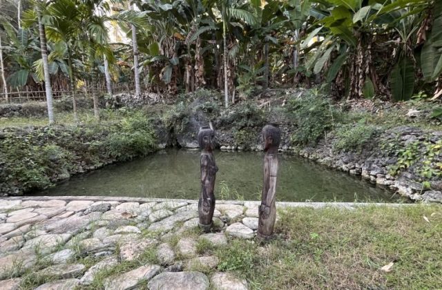 Two dark wooden statues of a male and female stand on a stone path beside a clear spring pool, surrounded by lush green tropical foliage.