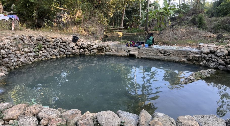 Two natural spring pools with stone walls are surrounded by lush green vegetation, as a person in a green shirt kneels by the far pool doing laundry.