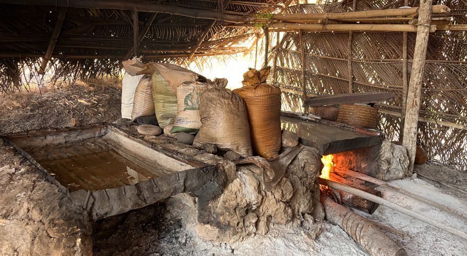 Inside a traditional Waiwuang salt-making hut, a fire intensely heats a stone structure with a water basin, while sacks of salt rest nearby.