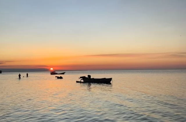 A vibrant orange and red sunset illuminates the calm waters of Walakiri Beach, where silhouetted figures wade and small boats float.