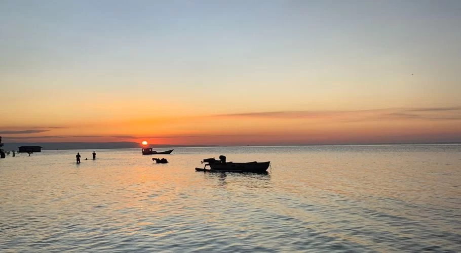 A vibrant orange and red sunset illuminates the calm waters of Walakiri Beach, where silhouetted figures wade and small boats float.