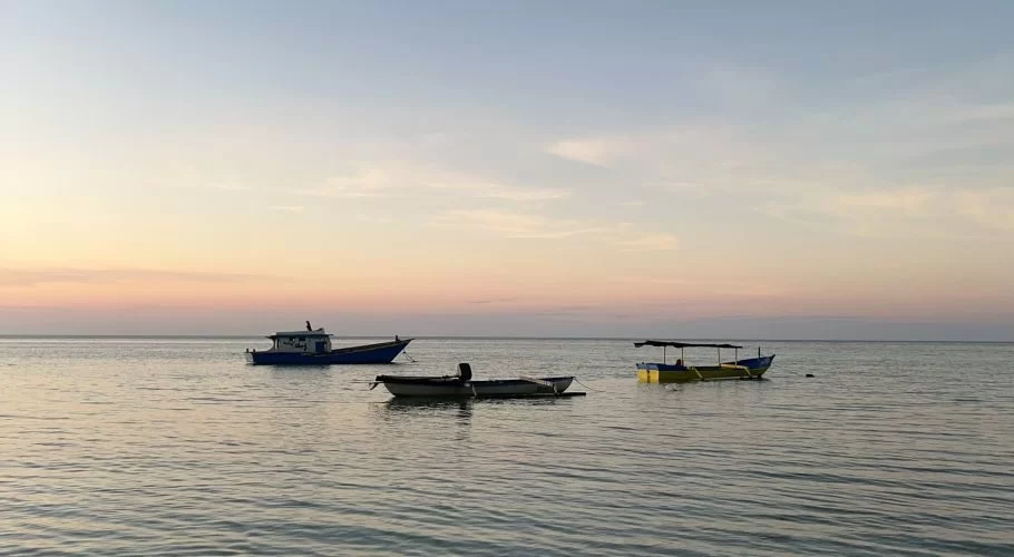 Three small boats, one blue, one white, and one yellow, float calmly on the serene, shallow waters of Walakiri Beach under a pastel sunset sky.