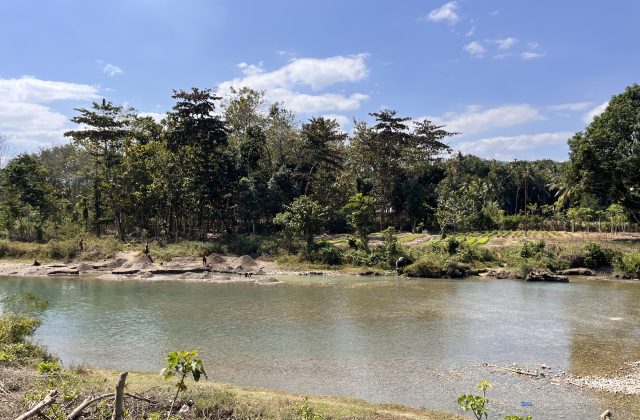 The wide Wanokaka River flows under a blue sky, with several people on its banks collecting stones amidst lush green trees and vegetation.