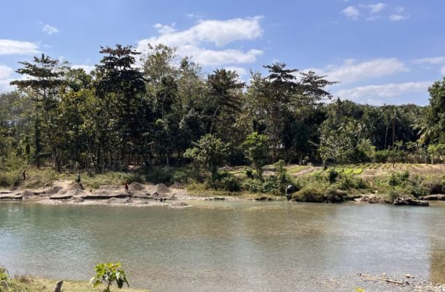 The wide Wanokaka River flows under a blue sky, with several people on its banks collecting stones amidst lush green trees and vegetation.
